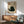May include: Framed artwork showing a black cat illustration with Japanese characters and red stamp seal, displayed above a wooden sideboard with white table lamp in a mid-century modern living room setting with mint green upholstered chair and round coffee table, all bathed in natural sunlight creating geometric shadows on white walls.