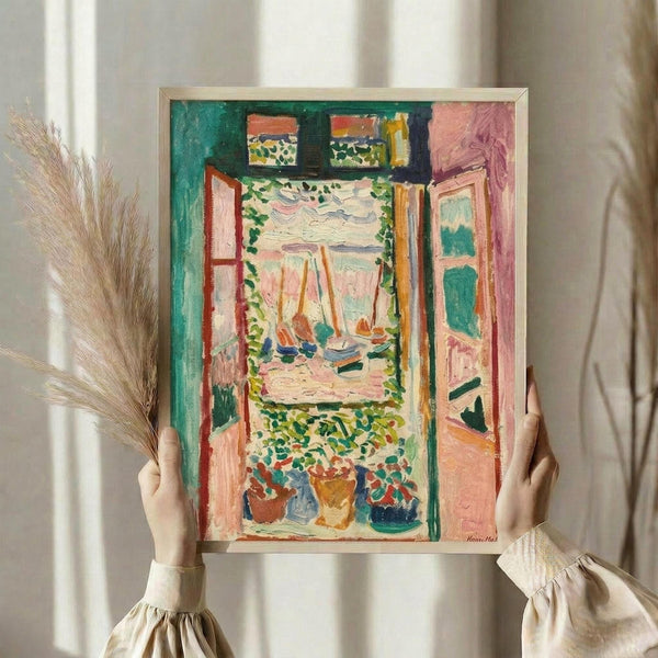 May include: Hands holding a framed colorful Fauve-style painting showing an open window view with sailboats on water, potted plants below, and vibrant pink, green, and orange brushstrokes. Person wears cream ruffle-sleeve top, holds dried wheat stems, against soft white curtained background.
