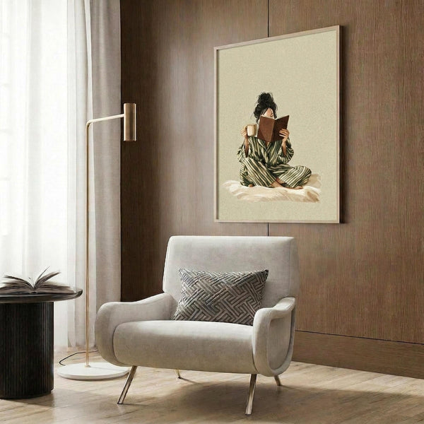 May include: Framed poster showing a woman in green striped pajamas sitting cross-legged on bed reading a book while holding coffee mug, displayed above cream upholstered chair with geometric pillow in modern room with wood panel walls, brass floor lamp, and side table with open book