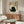 May include: Modern living room interior featuring a framed poster with Japanese calligraphy and a black ink-style cat illustration on cream textured background. The artwork hangs above a wood sideboard with sliding doors, flanked by a white curved chair, brass floor lamp, and white table lamp. An oval glass coffee table sits in the foreground with abstract blue and yellow artwork visible on its surface.