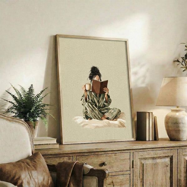 May include: Framed artwork displaying a woman in striped green pajamas sitting cross-legged on white bedding, reading a brown book while holding a mug, set against a cream background. The light wood frame rests on a rustic wooden console alongside books, a ceramic vase, and a fern plant. A beige upholstered chair with brown throw blanket is visible in the cozy, neutral-toned room setting.