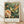 May include: Framed poster print leaning against white wall on light wood floor with natural shadows. Features vibrant hummingbird with yellow head, red throat, and green body hovering near large peach and cream colored flower surrounded by lush green foliage. Art displays rich botanical textile pattern in William Morris style with exhibition text below image in light wood frame.