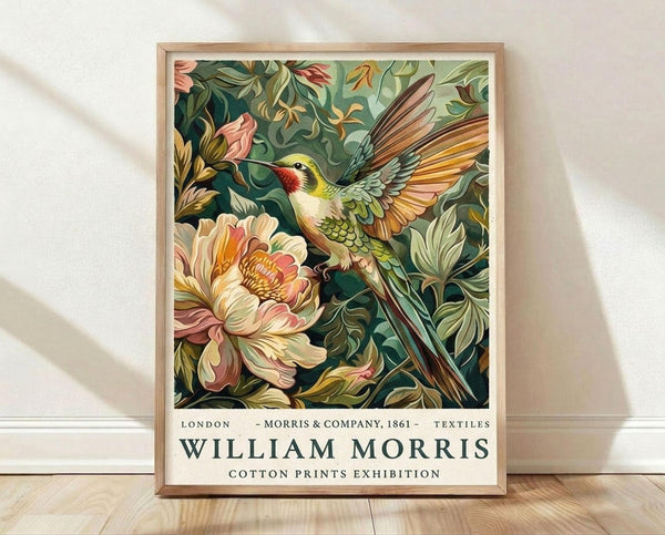 May include: Framed poster print leaning against white wall on light wood floor with natural shadows. Features vibrant hummingbird with yellow head, red throat, and green body hovering near large peach and cream colored flower surrounded by lush green foliage. Art displays rich botanical textile pattern in William Morris style with exhibition text below image in light wood frame.