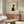 May include: Framed minimalist Japanese-style artwork featuring a black and white cat looking upward, displayed above a warm walnut wood credenza in a neutral living room. The print shows simple brushstroke style with the cat in profile against cream background. Room includes white curved chair, glass coffee table, and modern table lamp with white shade.