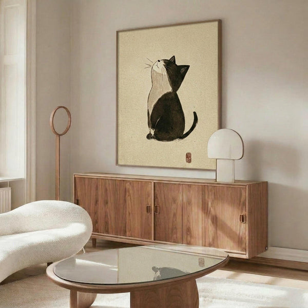 May include: Framed minimalist Japanese-style artwork featuring a black and white cat looking upward, displayed above a warm walnut wood credenza in a neutral living room. The print shows simple brushstroke style with the cat in profile against cream background. Room includes white curved chair, glass coffee table, and modern table lamp with white shade.