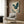 May include: Japanese-style artwork depicting a blue-green heron with outstretched wings soaring through swirling clouds, displayed in a light wooden frame on wood-paneled wall. Modern reading nook setting with cream upholstered armchair, geometric patterned pillow, brass floor lamp, and round side table with open book. Warm natural lighting from window with sheer curtains creates cozy atmosphere.