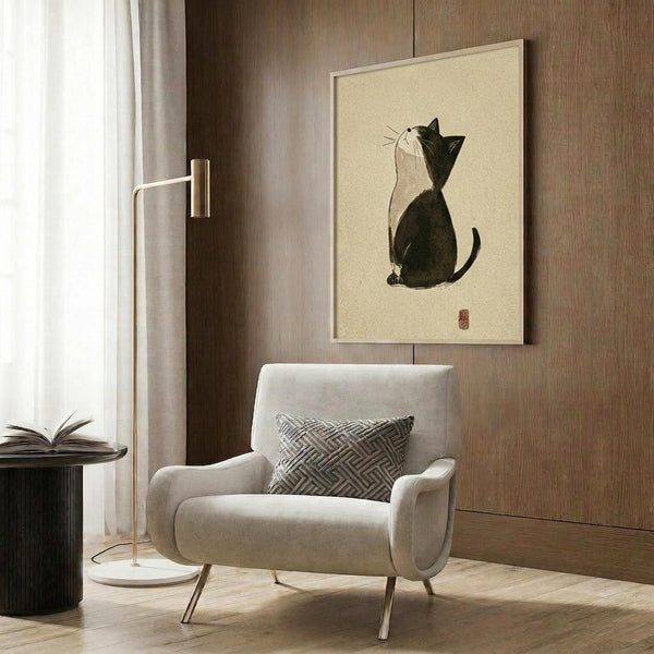 May include: Framed print showing a black and white cat sitting in profile, rendered in Japanese minimalist ink wash style on cream background with red seal stamp. Displayed on wood panel wall above beige upholstered chair with geometric patterned pillow, brass floor lamp, and dark round side table with open book in modern neutral-toned room.