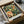 May include: Vintage-style botanical poster featuring colorful hummingbird with spread wings flying among lush green foliage and large pink peony flowers. Print shown in black frame inside cardboard shipping box with white tissue paper packaging on white fabric surface.