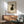 May include: Framed minimalist Japanese-style artwork featuring a black and white cat sitting and looking upward, displayed above a wooden sideboard with sliding doors. The print has a warm beige background with thin wooden frame. Scene shows mid-century modern furniture including a sage green upholstered armchair and white ceramic table lamp, with natural sunlight creating geometric shadows on white walls.
