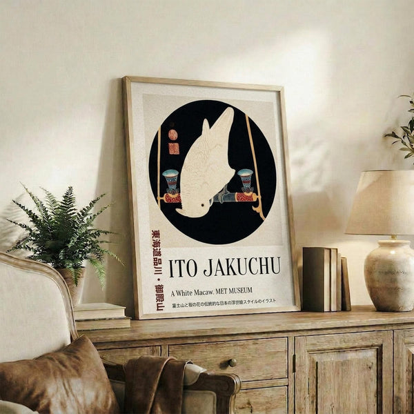 May include: Framed poster featuring a white parrot silhouette in black circle with decorative elements and "ITO JAKUCHU" text below, displayed on rustic wooden sideboard next to fern plant, books, ceramic vase, and beige upholstered chair in neutral-toned living room setting with natural lighting.