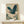 May include: Framed Japanese-style artwork featuring a blue-green heron with outstretched wings flying through swirling clouds and mist, displayed on a light wood console table next to a small white wrapped gift with greenery, against a cream-colored wall with natural lighting casting soft shadows.