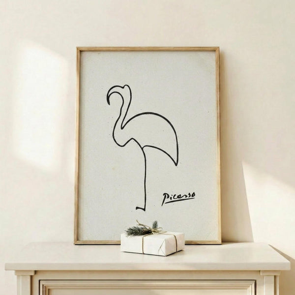 May include: Minimalist line drawing of a flamingo in black ink on white paper, displayed in a thin wooden frame on a light wood shelf against a cream wall. A small white gift box with twine and evergreen sprig sits beside the frame. The artwork features simple, flowing curves that form the flamingo's distinctive neck and body shape, with "Picasso" written in script below the drawing.