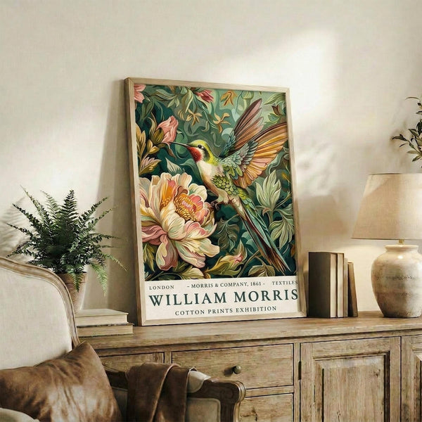 May include: Framed botanical poster featuring a vibrant hummingbird with spread wings hovering near a large pink peony flower, surrounded by lush green foliage in William Morris textile style. The print sits on a rustic wooden sideboard next to books, a ceramic vase, and fern plant in a bright living room setting with cream walls and beige furniture.