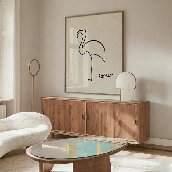 May include: Minimalist line drawing of a flamingo with Picasso signature displayed in thin wooden frame above walnut credenza in modern living room with cream walls, white sculptural chair, glass coffee table, and contemporary lighting fixtures