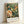 May include: Framed poster featuring a vibrant hummingbird with spread wings surrounded by lush botanical elements including large peony flowers and detailed foliage in rich greens, corals, and cream tones. The vintage-style illustration shows intricate feather details and decorative floral patterns. Text at bottom reads "London Morris & Company 1861 Textiles William Morris Cotton Prints Exhibition" in elegant typography. Light wood frame leans against white wall.