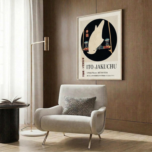 May include: A framed Japanese woodblock print featuring a white macaw in a black circle, displayed on a wood-paneled wall above a cream upholstered armchair. The poster shows traditional Japanese art style with the bird rendered in pale tones against dark background. Modern room setting includes geometric patterned pillow, brass floor lamp, and dark round side table with open book.