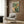 May include: Framed botanical artwork featuring a colorful hummingbird with spread wings among lush tropical flowers and foliage in greens, yellows, and coral tones. The print hangs on a wood-paneled wall above a cream upholstered armchair with geometric pillow. A brass floor lamp and side table with open book complete the warm, modern reading nook with natural lighting from nearby window.
