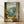 May include: Framed poster print leaning against white wall on light wood floor. Artwork shows Mediterranean coastal scene through open window with weathered shutters. Foreground features scattered yellow lemons, glass bottle with golden liquid, and lemon branch in vase on white cloth-covered windowsill. View reveals turquoise waters, hillside village with white buildings, and mountain backdrop under cloudy sky. Warm natural lighting creates shadows on wall.
