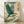 May include: Framed poster print featuring a blue-green heron in flight with outstretched wings, surrounded by stylized cream and teal clouds and waves in traditional Japanese art style. The bird has a long pointed beak and reddish legs. A red Japanese character seal appears in the upper right corner. The artwork is displayed in a light wooden frame, leaning against a white wall on light wood flooring with soft natural lighting casting shadows.
