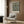 May include: Framed botanical poster with intricate floral pattern in black, gold and green tones hanging on wood-paneled wall above beige upholstered armchair with geometric patterned pillow. Modern brass floor lamp and round black side table with open book complete the elegant reading nook setting with natural lighting from nearby window.