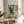 May include: Framed Japanese-style artwork of a blue-green heron in flight with outstretched wings, surrounded by swirling cream and teal clouds and waves, displayed on a light wood round side table next to a beige upholstered chair with white throw pillow, plant leaves visible in corner, natural lighting creating soft shadows on white wall