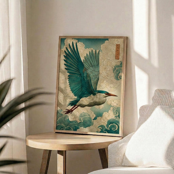 May include: Framed Japanese-style artwork of a blue-green heron in flight with outstretched wings, surrounded by swirling cream and teal clouds and waves, displayed on a light wood round side table next to a beige upholstered chair with white throw pillow, plant leaves visible in corner, natural lighting creating soft shadows on white wall