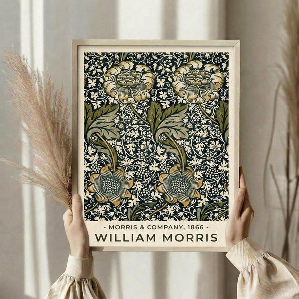 May include: Hands holding a framed botanical print featuring William Morris's ornate floral pattern in navy blue, sage green, and cream tones. The vintage-style design shows detailed flowers, leaves, and decorative scrollwork. Light wood frame against neutral curtain backdrop with dried pampas grass visible on left side.