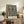 May include: Framed botanical art print with intricate black floral pattern featuring cream and green blooms on dark background, displayed on rustic wooden sideboard alongside fern plant, ceramic vases, and leather armchair in neutral-toned living room setting