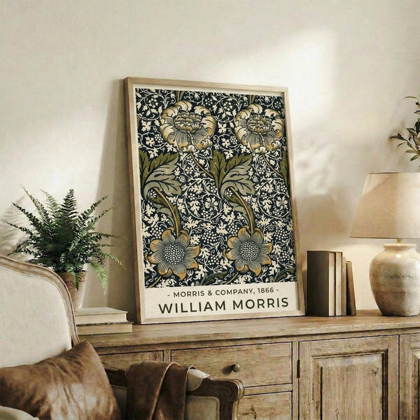 May include: Framed botanical art print with intricate black floral pattern featuring cream and green blooms on dark background, displayed on rustic wooden sideboard alongside fern plant, ceramic vases, and leather armchair in neutral-toned living room setting