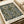 Vintage William Morris textile design print featuring intricate floral pattern with large golden blooms, green leaves, and ornate scrollwork on dark navy background. The botanical wallpaper design shows detailed linework and rich colors typical of Arts and Crafts movement. Print appears mounted or framed with cream border, photographed alongside neutral wrapping paper and ribbon, showing fine detail and craftsmanship of the decorative pattern.