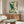 May include: Japanese-style artwork featuring a blue-green heron with outstretched wings flying through stylized cream and teal clouds, displayed above a mid-century modern walnut credenza in a minimalist living room with white curved chair, oval glass coffee table, and warm natural lighting