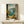 May include: Framed artwork displaying an open window view of the Amalfi Coast with turquoise waters, mountainous coastline, and a windowsill arrangement of fresh lemons, olive oil bottles, and Mediterranean plants. The painting features realistic brushwork in blues, yellows, and earth tones. A small white wrapped package sits beneath the wooden-framed print on a light wooden shelf against a cream wall.