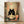 May include: A framed poster print displayed on wooden flooring against a white wall. The artwork features a black cat with large round white eyes sitting behind a bowl of ramen noodles, with chopsticks and a bottle nearby. The background shows a collage of vintage Japanese food advertisements and signs in muted colors including red, beige, and green. The print appears in a natural wood frame and casts soft shadows on the wall.