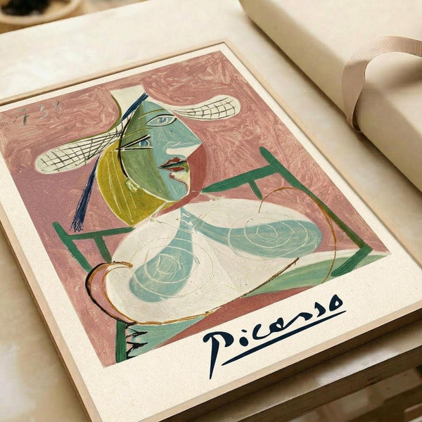 Abstract cubist-style artwork featuring a figure with green and yellow tones, geometric shapes, and flowing lines on a pink background. The piece shows characteristic angular features with multiple eyes and curved elements, signed "Picasso" in black ink at the bottom. The print is displayed on cream-colored paper alongside rolled paper tubes on a neutral surface.