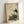 May include: Framed vintage Japanese-style artwork showing a white stork with black wing feathers standing on a rock next to a brown turtle, surrounded by green foliage. The print features traditional Asian calligraphy on aged beige paper with red seal stamps. The simple wooden frame leans against a white wall, casting a soft shadow.