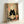 May include: Framed poster print leaning against white wall showing adorable black cat with large round eyes sitting behind bowl of ramen noodles with chopsticks. Background features collage of vintage Japanese restaurant signs and text in muted colors. Light wood frame, cozy kawaii art style with warm beige and red tones throughout the illustrated design.