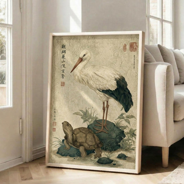 May include: Framed vintage-style Japanese artwork featuring a white stork with dark wing tips standing beside a brown turtle on blue rocks with green plants. The print has aged beige background with traditional Asian calligraphy and red seals. Light wood frame leans against white wall next to beige sectional sofa in bright modern living room with large windows.