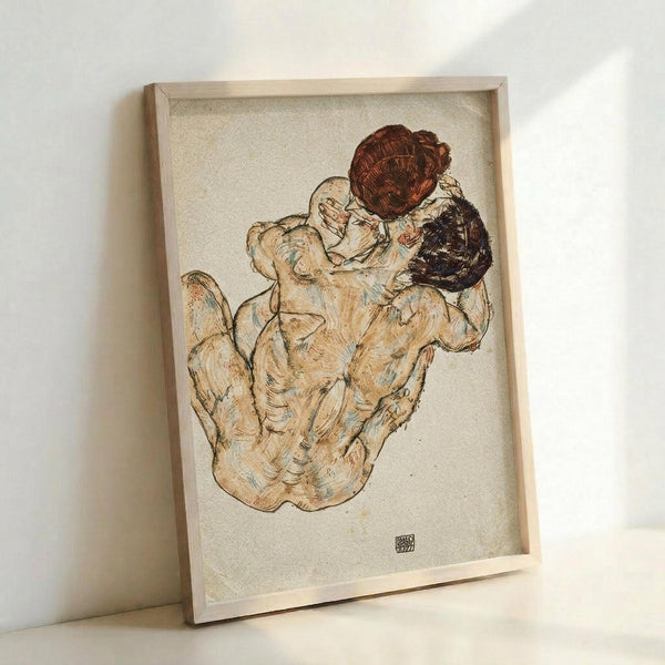 Framed art print showing an intimate sketch of two figures embracing in warm earth tones of brown, beige and cream. The drawing depicts intertwined bodies rendered in expressive line work with watercolor washes. The artwork is displayed in a light wooden frame against a white wall, with the artist's signature monogram visible in the lower right corner of the composition.