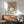 May include: Framed Picasso-style cubist portrait poster displayed above a mid-century modern wooden credenza with table lamp. Green upholstered armchair with wooden frame sits nearby on light wooden floors. Artwork features abstract female figure in pink, green, and yellow tones with geometric styling. Natural sunlight creates dramatic shadows across white walls in minimalist interior setting.
