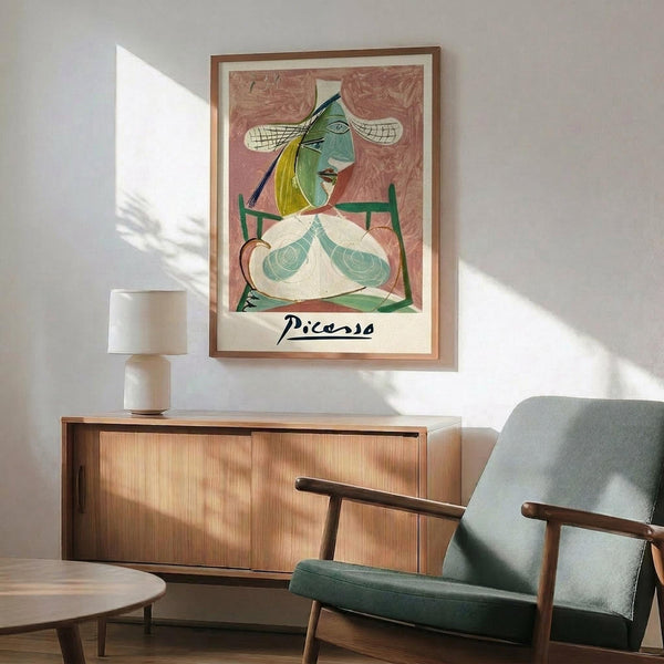 May include: Framed Picasso-style cubist portrait poster displayed above a mid-century modern wooden credenza with table lamp. Green upholstered armchair with wooden frame sits nearby on light wooden floors. Artwork features abstract female figure in pink, green, and yellow tones with geometric styling. Natural sunlight creates dramatic shadows across white walls in minimalist interior setting.
