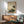 May include: Traditional Japanese-style artwork depicting a white stork with black wing tips standing beside a brown turtle, surrounded by green vegetation and rocks. The vintage print features Asian calligraphy and red seals on aged beige paper with a thin brown frame. Displayed above a wooden sideboard with sliding doors next to a gray upholstered chair with wooden arms in a modern living room with white walls and natural lighting.