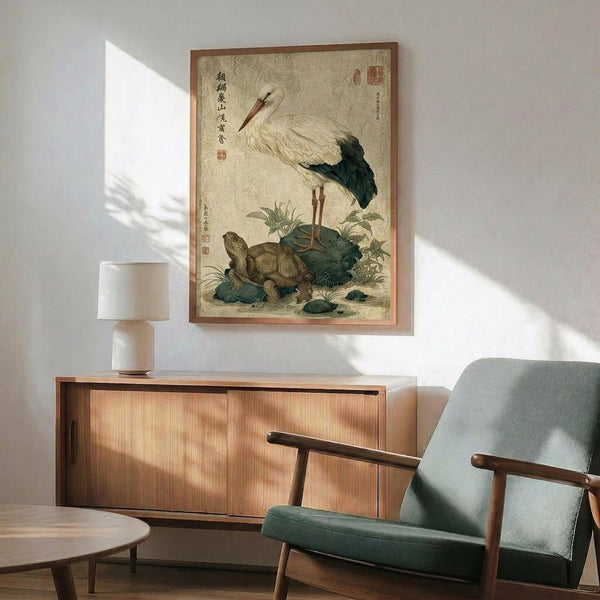 May include: Traditional Japanese-style artwork depicting a white stork with black wing tips standing beside a brown turtle, surrounded by green vegetation and rocks. The vintage print features Asian calligraphy and red seals on aged beige paper with a thin brown frame. Displayed above a wooden sideboard with sliding doors next to a gray upholstered chair with wooden arms in a modern living room with white walls and natural lighting.