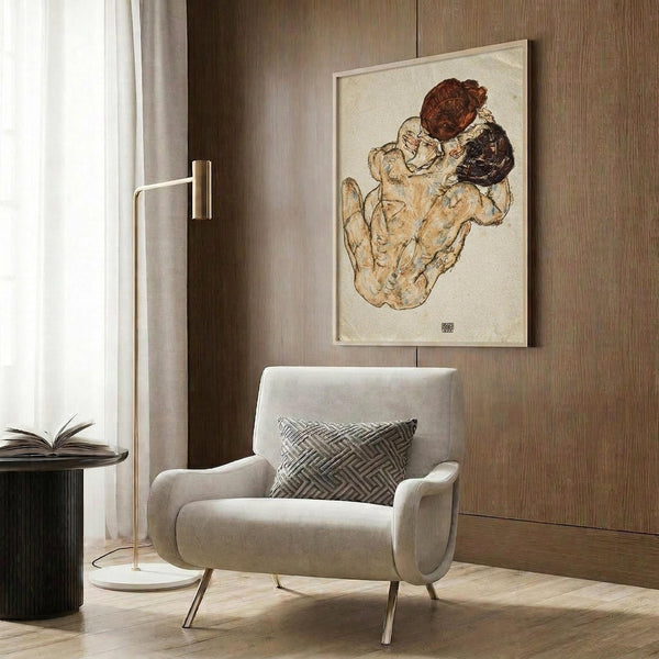 May include: Framed reproduction of Schiele's expressive line drawing showing two embracing figures with auburn and dark hair rendered in warm earth tones, displayed on wood-paneled wall above modern beige armchair with geometric pillow, next to brass floor lamp and dark side table with open book.