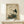May include: Traditional Japanese-style artwork in natural wood frame showing white stork with dark wing tips standing beside brown turtle on rocks with green foliage. Print features aged beige background with black Japanese calligraphy and red seal stamps. Frame displayed on light wood surface next to small white wrapped gift against cream wall.