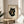 May include: Framed artwork featuring a black cat with large golden eyes eating ramen noodles with chopsticks from a dark bowl, rendered in Japanese ukiyo-e woodblock print style on aged cream paper with red seal stamp. Wooden frame sits on round wooden side table next to beige upholstered chair with decorative pillow, green plant leaves visible, soft natural lighting creates shadows on neutral wall.