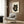 May include: A framed poster featuring a black cat with large golden eyes eating ramen noodles with chopsticks from a dark bowl, rendered in traditional Japanese ukiyo-e art style on cream-colored paper. The artwork hangs on a wood-paneled wall above a beige upholstered armchair with geometric throw pillow, beside a brass floor lamp and dark round side table with an open book.