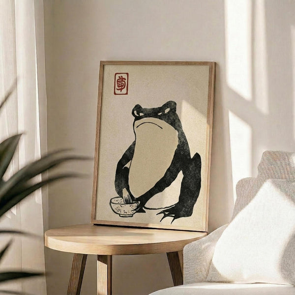 May include: Japanese woodblock-style print in thin wooden frame showing dark green frog sitting with a small bowl, featuring red square seal stamp at top, displayed on round wooden side table next to beige upholstered chair with white pillow, soft natural lighting creating shadows on white wall background