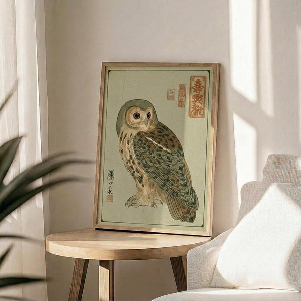 May include: Framed vintage Japanese woodblock print featuring a detailed owl with brown and green feathers, surrounded by traditional calligraphy characters on pale green background, displayed on round wooden side table next to beige upholstered chair and plant leaves in bright, naturally lit room with white walls.