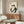 May include: Minimalist living room with cream walls featuring a framed Japanese-style artwork of a dark green frog sitting with a bowl. The print hangs above a walnut wood credenza with sliding doors. Modern white curved chair and oval glass coffee table with blue abstract pattern complete the scene. Warm natural lighting creates shadows on the furniture.