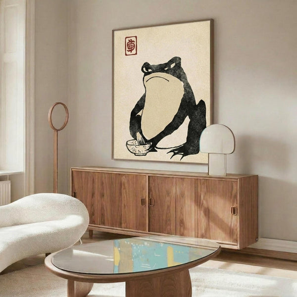 May include: Minimalist living room with cream walls featuring a framed Japanese-style artwork of a dark green frog sitting with a bowl. The print hangs above a walnut wood credenza with sliding doors. Modern white curved chair and oval glass coffee table with blue abstract pattern complete the scene. Warm natural lighting creates shadows on the furniture.